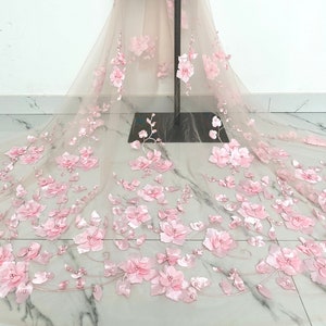 Puede incluir: Tela de encaje floral rosa con flores 3D y detalles de perlas. El encaje es delicado y transparente, y las flores son suaves y femeninas.