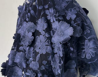 May include: Close-up of navy blue floral lace fabric. The fabric features embroidered flowers in a lighter shade of blue, creating a three-dimensional effect. The delicate design is visible against a dark background, showcasing the intricate details.