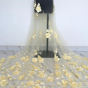 Peut inclure: Un tissu jaune pâle transparent orné de nombreuses fleurs jaune clair en trois dimensions. Le tissu est drapé sur un mannequin noir, avec des ornements floraux en cascade et traînant sur le sol.