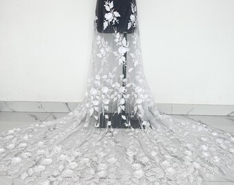May include: White floral lace fabric with 3D flower appliques. The delicate fabric is draped over a mannequin, showcasing the intricate floral design. The flowers are white and the background is a sheer, light color.