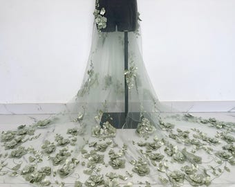May include: A sheer, light green fabric adorned with three-dimensional, sage green floral appliqués. The fabric is draped over a dress form, with the floral embellishments cascading down the front. The flowers vary in size and are clustered throughout the fabric.