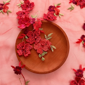 May include: Close-up of red floral embroidery on sheer pink fabric. The embroidery features detailed red flowers with green leaves, arranged around a wooden plate. The overall effect is delicate and decorative.