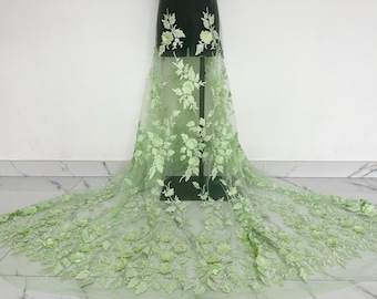 May include: Light green floral lace fabric with 3D flower appliques. The fabric is displayed on a dress form, showcasing the intricate design. The flowers are a lighter shade of green, creating a beautiful contrast.