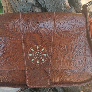 Puede incluir: Un bolso de hombro de cuero marrón con un intrincado patrón floral en relieve en toda la superficie. La solapa frontal incluye un diseño circular central con pequeños detalles en verde claro y una correa de cuero a juego con herrajes metálicos.