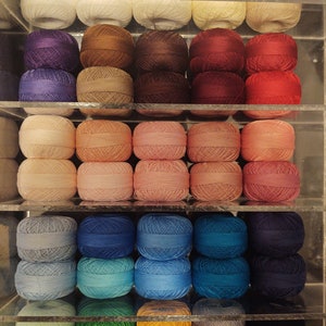 May include: A collection of yarn balls in various colors, including white, purple, brown, pink, blue, green, orange, and gray, are displayed on shelves in a clear acrylic storage container.