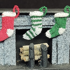 May include: A miniature fireplace with three knitted stockings hanging from the mantel. The stockings are red, green and white with a white trim. The fireplace is made of gray cardboard and has a small log fire inside.