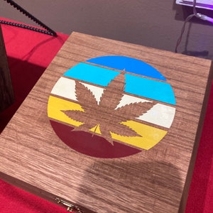 May include: A wooden box with a colorful, retro-style design featuring a marijuana leaf. The design includes a circle with stripes of blue, turquoise, white, yellow, and red.