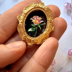 Pintura al óleo en miniatura de una rosa color crema sobre fondo oscuro, con marco dorado ovalado e imán, ideal para decorar casas de muñecas. Arte floral.