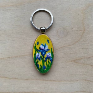 May include: A yellow oval wooden keychain with a painted image of three blue and white iris flowers with green leaves.