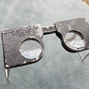 May include: A black metal magnifying glass with two lenses. The magnifying glass has a metal frame and is adjustable. The text "ABRAMS INSTRUMENT CO. LANSING, MICH. U.S.A." is engraved on the frame.