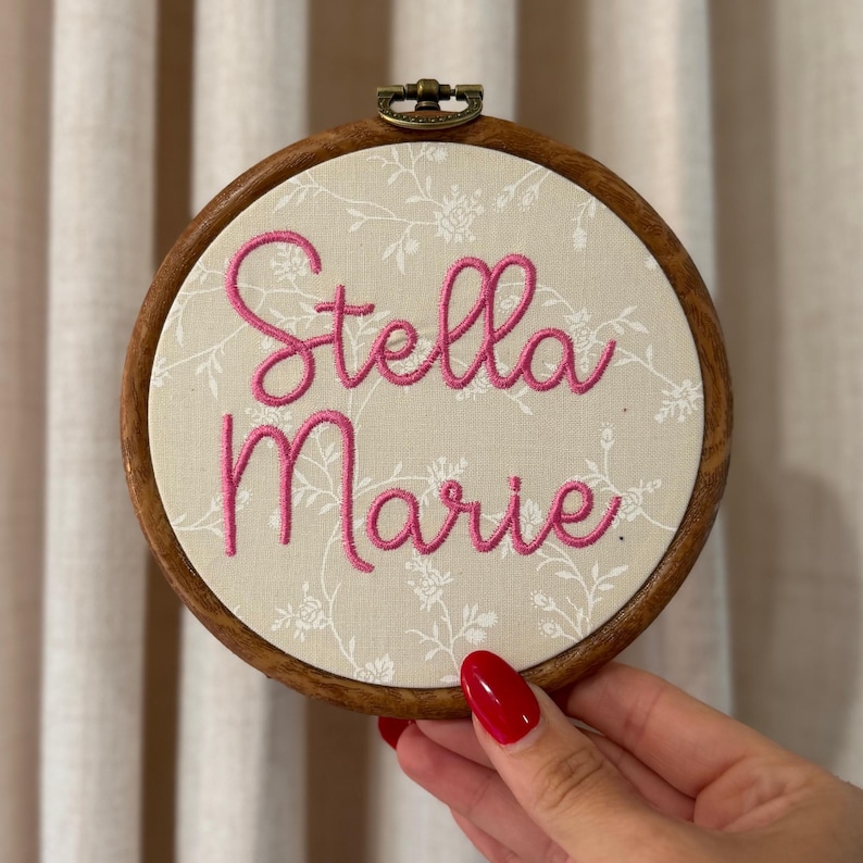 May include: A circular wooden embroidery hoop containing cream-colored fabric with a white floral pattern. The name Stella Marie is embroidered in the center using bright pink thread in a cursive script font. The hoop features a metal tension screw.