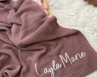 May include: A dusty rose knitted baby blanket featuring a textured garter stitch pattern and a wide ribbed border. The name Layla Marie is embroidered in white cursive script on the lower corner of the blanket, which is draped over a baby.