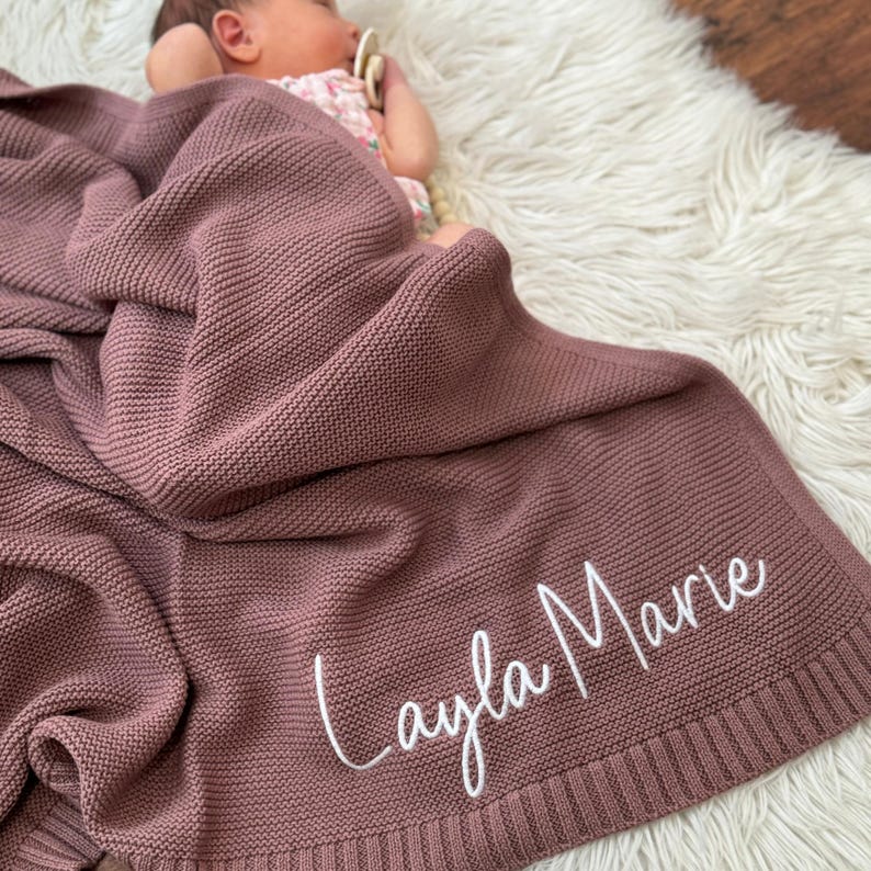 Pu&ograve; includere: Una coperta per neonati lavorata a maglia color rosa polvere con un motivo a legaccio testurizzato e un ampio bordo a coste. Il nome Layla Marie &egrave; ricamato in corsivo bianco sull'angolo inferiore della coperta, drappeggiata su un neonato.