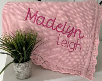 Può includere: Una coperta lavorata a maglia rosa chiaro con un bordo decorativo smerlato e il nome Madelyn Leigh ricamato in corsivo di una tonalità di rosa più scura sulla superficie anteriore del tessuto.