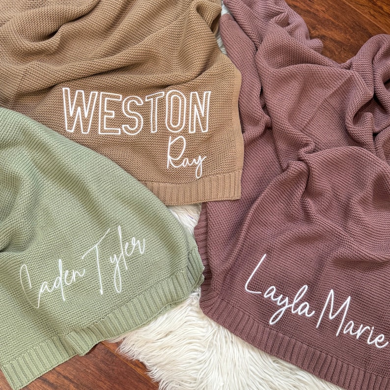 Pu&ograve; includere: Tre coperte lavorate a maglia nei colori marrone chiaro, verde salvia e malva. La coperta marrone chiaro presenta il testo bianco 'WESTON Ray', quella verde salvia il testo 'Caden Tyler' e quella malva il testo 'Layla Marie'.