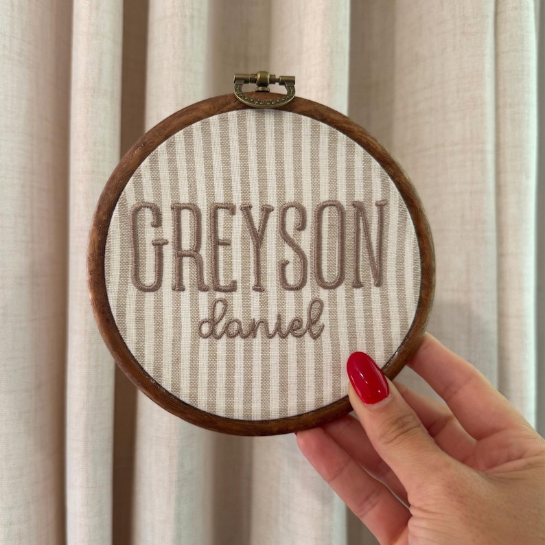 May include: A circular wooden embroidery hoop framing beige and white striped fabric. The name GREYSON is embroidered in large, dark brown capital letters, with the name daniel embroidered below in a smaller, dark brown cursive script font.