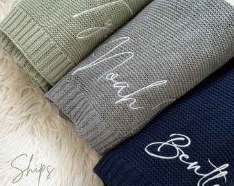 May include: Three folded knit blankets in sage green, gray, and navy blue, each embroidered with a name in white script. The blankets are arranged on a fluffy white surface. The names are: Taylor, Noah, and Bentley.