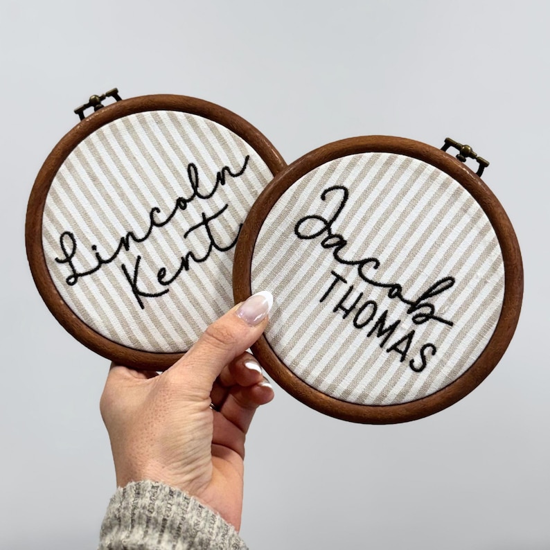 May include: Two circular wooden embroidery hoops containing beige and white striped fabric. The left hoop features the black embroidered text Lincoln Kent, and the right hoop features the black embroidered text Jacob THOMAS in a mix of script and block fonts.