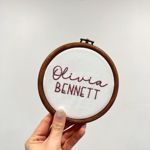 May include: A circular wooden embroidery hoop containing white fabric with the name Olivia BENNETT stitched in dark maroon thread. The top of the hoop features a small metal tightening screw mechanism for securing the fabric in place.