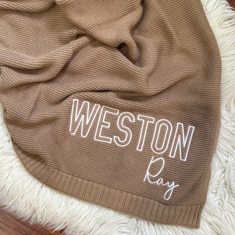 Pu&ograve; includere: Una coperta lavorata a maglia color marrone chiaro con il nome WESTON Ray ricamato in filo bianco. La coperta presenta un motivo a legaccio testurizzato e un bordo a coste sul fondo, creando un netto contrasto tra il tessuto e il testo.