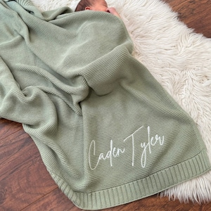 Pu&ograve; includere: Una coperta per neonati in maglia color verde salvia con un motivo a legaccio testurizzato e un bordo a coste. Il nome Caden Tyler &egrave; ricamato in corsivo bianco nell'angolo in basso a destra della coperta appoggiata su una superficie.