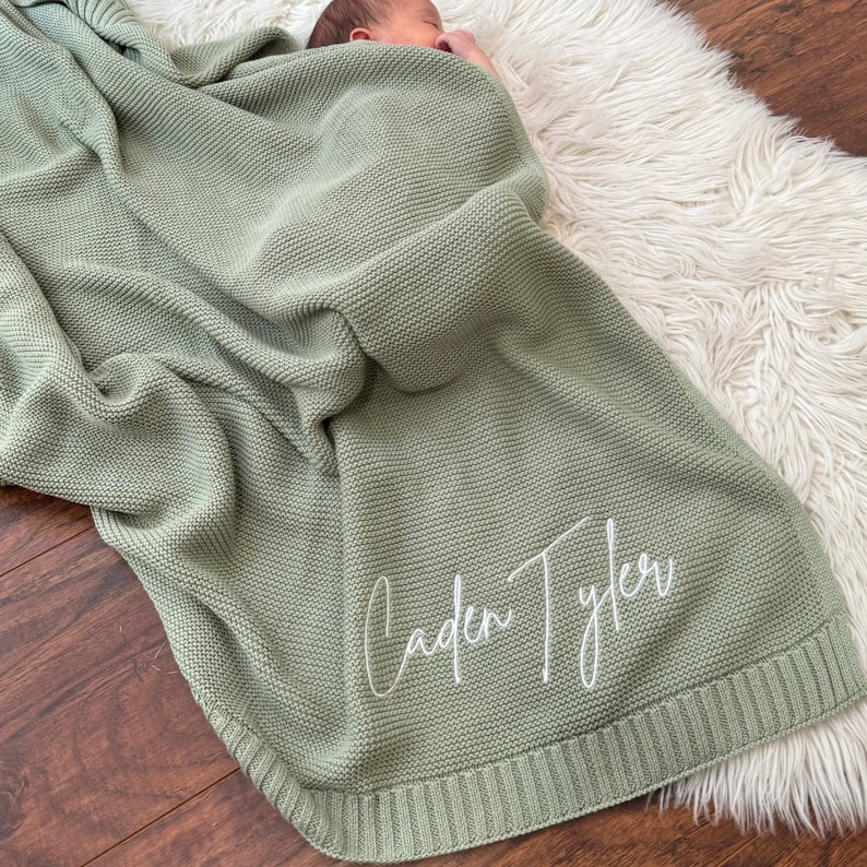 Pu&ograve; includere: Una coperta per neonati in maglia color verde salvia con un motivo a legaccio testurizzato e un bordo a coste. Il nome Caden Tyler &egrave; ricamato in corsivo bianco nell'angolo in basso a destra della coperta appoggiata su una superficie.
