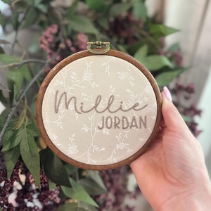 May include: A circular wooden embroidery hoop containing beige fabric with a white floral pattern. The name Millie Jordan is embroidered in brown thread across the center. The hoop features a small metal hanging hardware at the top.