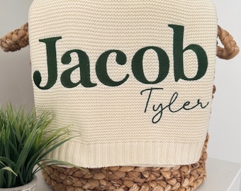 May include: Cream-colored knitted blanket with the name "Jacob" in dark green block letters and the name "Tyler" in a cursive font. The blanket is folded inside a woven basket with handles.