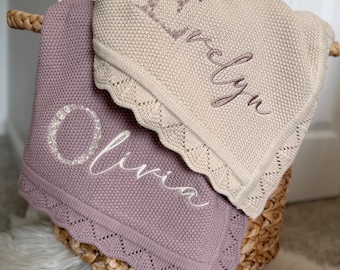 May include: Two personalized baby blankets in a wicker basket. One is a light purple color with the name "Olivia" embroidered in white. The other is a tan color with the name "Evelyn" embroidered in a matching tan color. Both blankets have a scalloped edge.