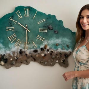 Puede incluir: Un gran reloj de pared con un diseño temático del océano. La esfera del reloj presenta un fondo de resina verde azulado con un efecto de olas y arena, y piedras incrustadas. Los números romanos dorados marcan las horas. El reloj mide aproximadamente 76 cm de diámetro.