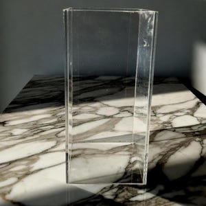 May include: A clear, rectangular acrylic display case with a faceted design. The case is empty and sits on a marble-patterned surface. The lighting creates shadows, highlighting the case's shape and transparency.