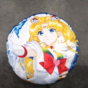 Könnte beinhalten: Rundes Kissen mit einer farbenfrohen Anime-Illustration. Das Design zeigt eine Figur mit blonden Haaren, blauen Augen und einer großen roten Schleife. Das Kissen hat einen weißen und blauen Rand und ist wahrscheinlich aus weichem Stoff.