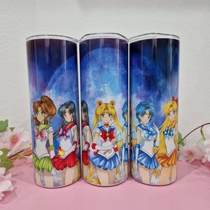 Könnte beinhalten: Drei zylindrische Becher mit einem Anime-Design. Jeder Becher zeigt einen anderen Charakter in einer Matrosenuniform vor dem Hintergrund eines blauen Nachthimmels mit einem großen Mond. Die Charaktere haben verschiedene Haarfarben und Outfits.
