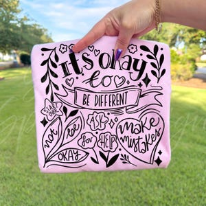 Op de afbeelding: Een lichtroze t-shirt met zwarte tekst en illustraties. De tekst luidt "It's okay to be different" en andere bemoedigende zinnen. Het ontwerp bevat harten, bloemen en bladeren, wat een positieve boodschap creëert.