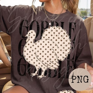 May include: A dark gray short-sleeve t-shirt featuring a large graphic of a turkey with a cream-colored body and brown polka dots. The background of the graphic displays the word GOBBLE repeated in large, bold, black, distressed-style lettering.