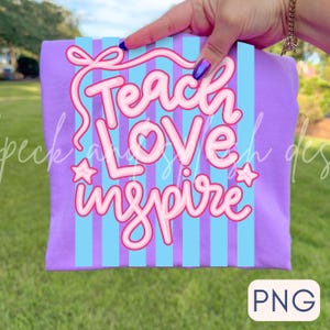 May include: A purple t-shirt with a design that says "Teach Love Inspire" in pink lettering. The design includes a bow, stars, and vertical blue stripes. The shirt is held up by a hand against a green grass background.