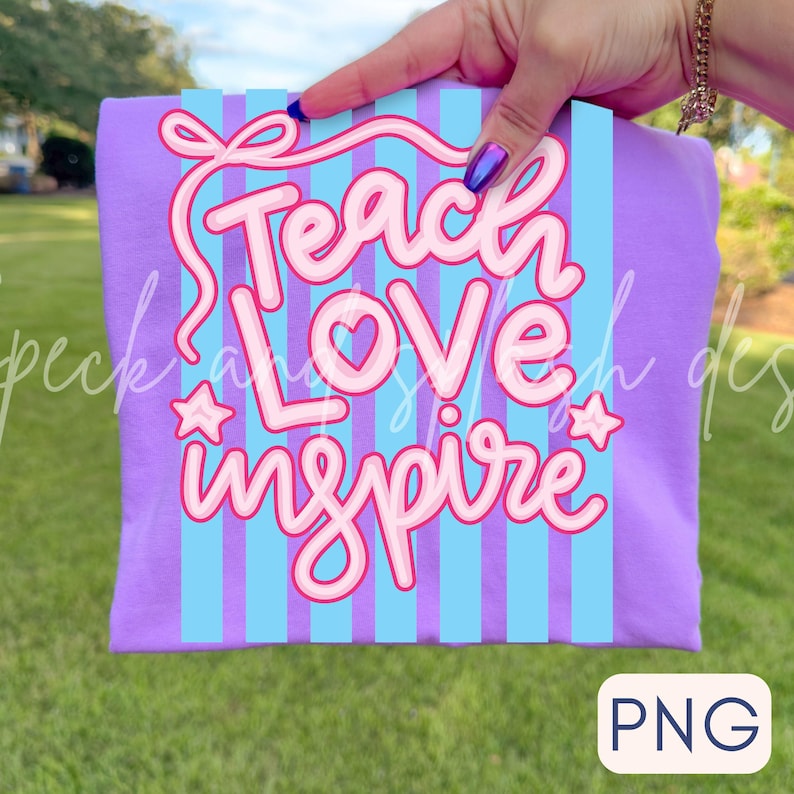 May include: A purple t-shirt with a design that says "Teach Love Inspire" in pink lettering. The design includes a bow, stars, and vertical blue stripes. The shirt is held up by a hand against a green grass background.
