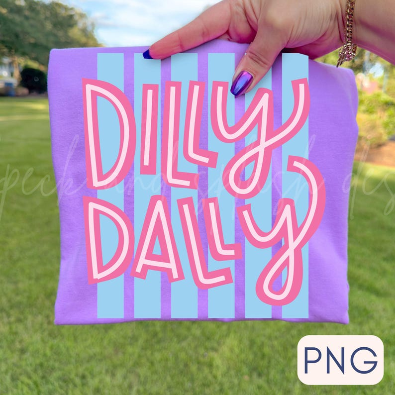 Puede incluir: Una camiseta de color lila con las palabras "Dilly Dally" en una fuente juguetona, rosa y blanca. El texto est&aacute; sobre un fondo a rayas azul claro. La camiseta se sostiene sobre un fondo de hierba verde.