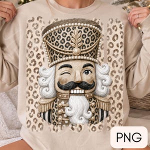Può includere: Una felpa beige con la grafica di uno schiaccianoci che fa l'occhiolino, con una stampa leopardata sul cappello e sull'uniforme. Lo schiaccianoci ha barba, baffi e capelli bianchi, con dettagli dorati e perle sul cappello.