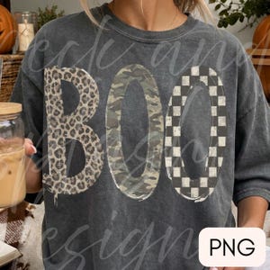 May include: A charcoal gray short-sleeve t-shirt featuring the word BOO printed in large letters. The letter B has a leopard print pattern, the letter O has a camouflage pattern, and the second O has a black and white checkered pattern.