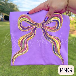 May include: A lilac-colored t-shirt featuring a large, colorful bow design. The bow has a rainbow of colors and is outlined with gold glitter. The shirt is held up by a hand, and the background is a grassy area.