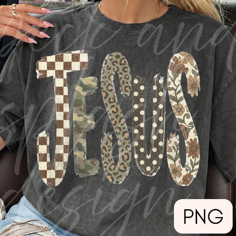 May include: A charcoal gray crewneck t-shirt featuring the word JESUS in large, stylized letters. Each letter has a unique pattern: a brown and white checkerboard, green camouflage, brown leopard print, white polka dots, and a floral design.