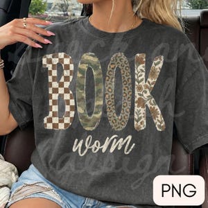 May include: A charcoal gray short-sleeve t-shirt featuring the word BOOK in large block letters with varied patterns: brown and white checkerboard, green camouflage, brown leopard print, and a floral design, with the word worm in white script.