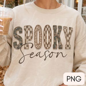May include: A beige crewneck sweatshirt featuring the word SPOOKY in large block letters with varied patterns including camouflage, checkerboard, and leopard print. Below, the word Season is written in a dark brown cursive script font.