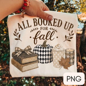 May include: A cream-colored t-shirt featuring the brown text ALL BOOKED UP FOR fall. The graphic includes a stack of two books with a plaid bow, a black and white diamond-patterned pumpkin, a polka-dot cup with whipped cream, and a plaid candle.