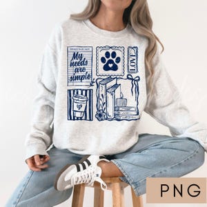 May include: A light gray sweatshirt with a navy blue graphic design. The design includes the text "My needs are simple" along with images of a coffee cup, books, a paw print, and the word "LOVE". The sweatshirt is a casual, comfortable style.
