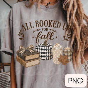 May include: A grey t-shirt featuring the brown text ALL BOOKED UP FOR fall, accompanied by a stack of two books with a plaid bow, a black and white checkered pumpkin, a cup of coffee with whipped cream, a lit candle, and a brown leaf.