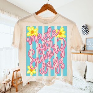 May include: Cream-colored t-shirt with the phrase "Made for Sunny Days" in pink script, set against a blue and white striped background. Yellow flower graphics accent the design. The shirt is hanging on a wooden hanger.