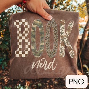 Op de afbeelding: Een bruin t-shirt met het woord BOOK in grote blokletters, elk met een ander patroon: bruin en wit dambord, groen camouflage, luipaardprint en een bloemmotief. Het woord nerd staat eronder gedrukt in wit cursief schrift.