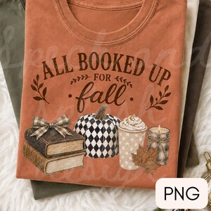 May include: A rust-colored t-shirt featuring the text ALL BOOKED UP FOR fall in dark brown lettering. The graphic includes a stack of two books with a plaid bow, a black and white checkered pumpkin, a polka-dot mug with foam, and a candle.